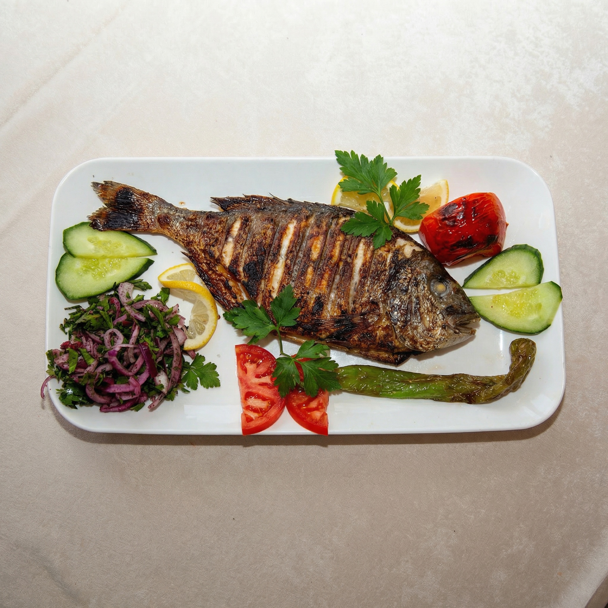 Servierplatte mit einer gegrillten Dorade, garniert mit Gurkenscheiben, Zwiebelsalat, Tomaten und Peperoni.