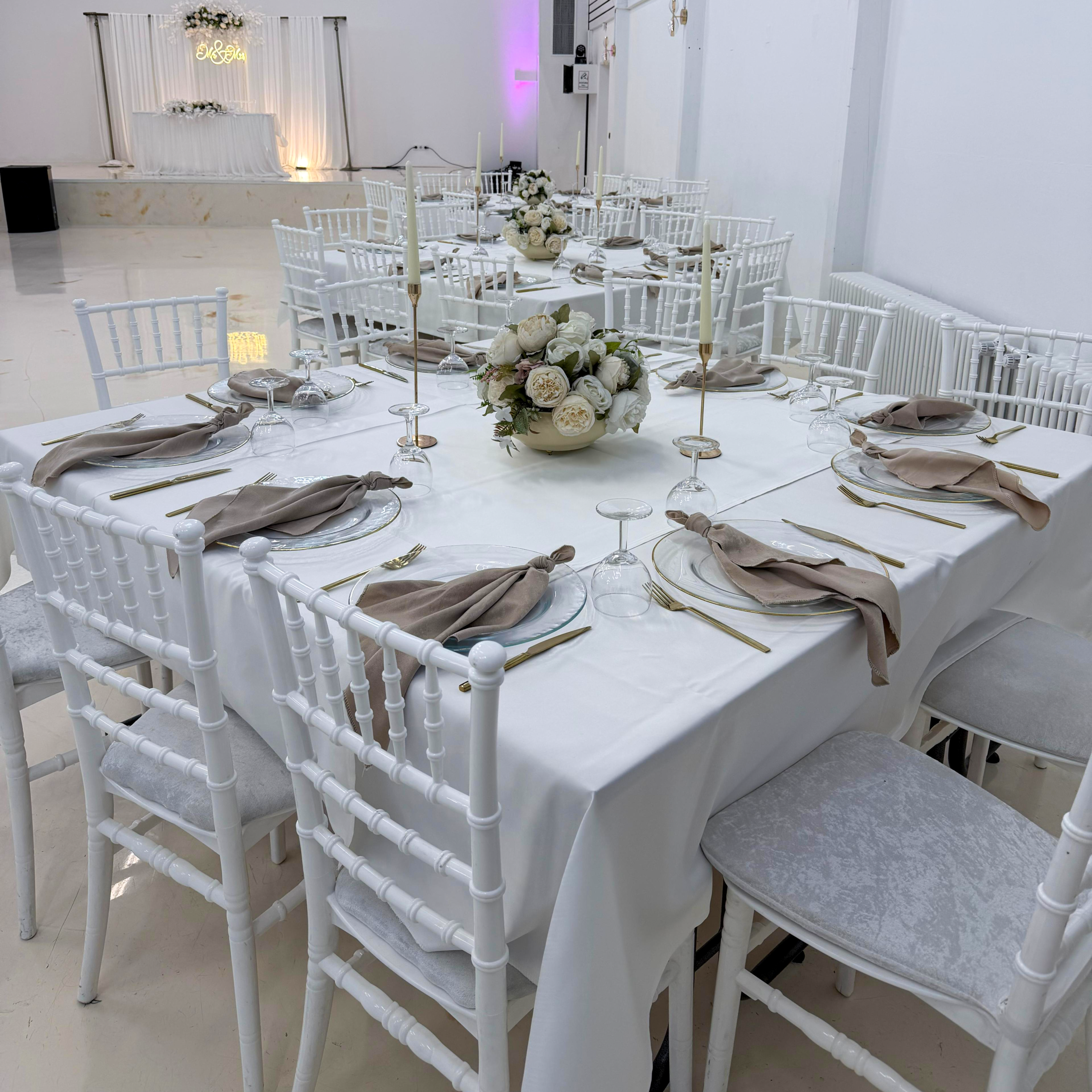Nahaufnahme einer quadratischen Festtafel mit weißen Stühlen, Stoffservietten und Blumen-Centerpiece.
