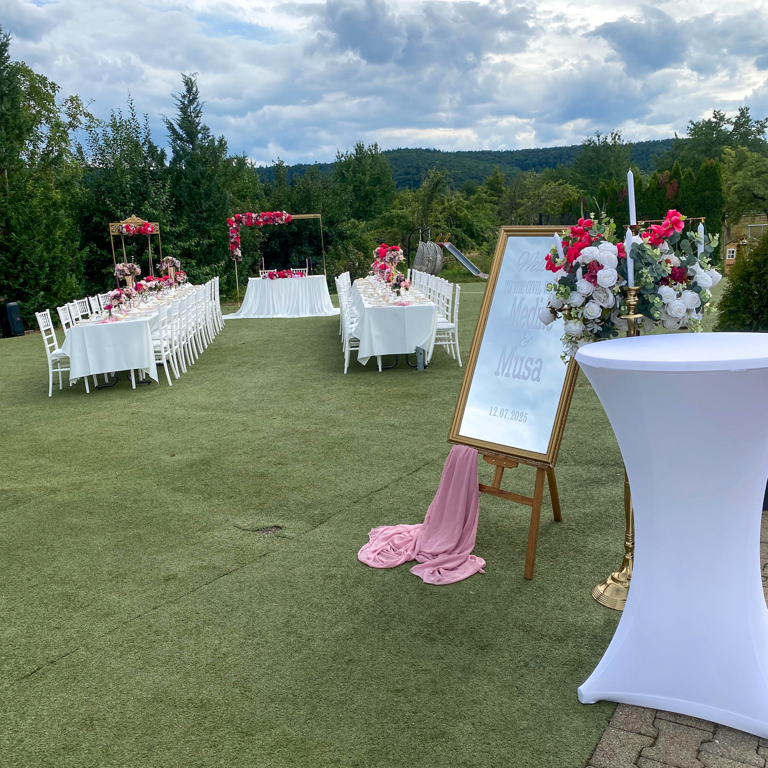Außenbereich mit weiß gedeckten Tischen auf Grünfläche und Willkommensschild für das Brautpaar.