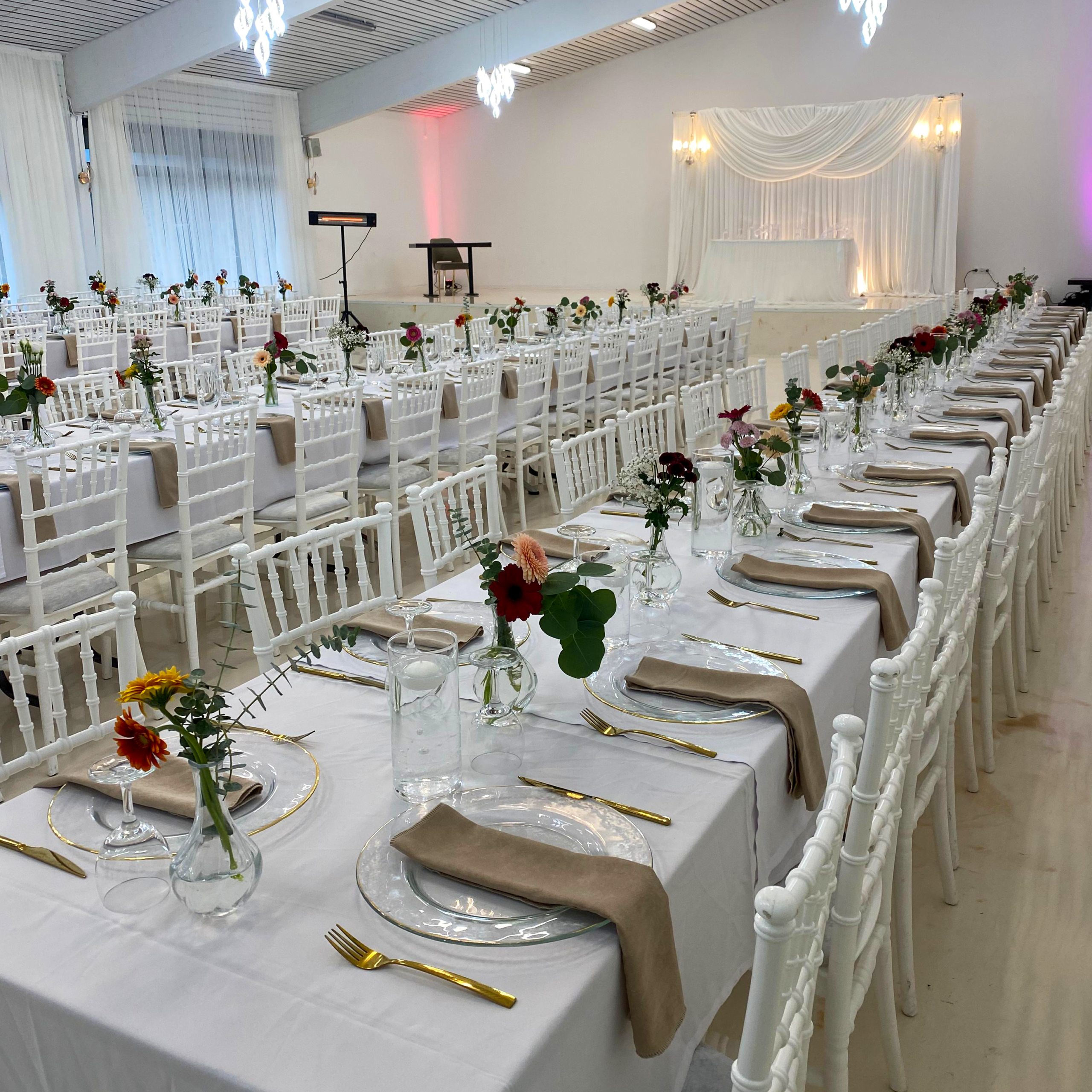 Lange Festtafeln mit bunten Wildblumen, beigen Servietten und goldenem Besteck im Festsaal.